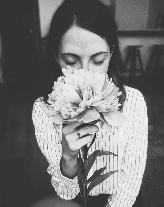 A black and white portrait of a woman with closed eyes, smelling a peony flower indoors.